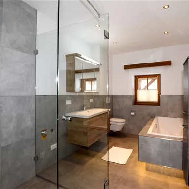 A modern bathroom with gray tiles and a large glass shower. There is a bathtub, a wash area, and a wooden cabinet.