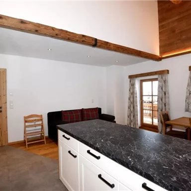 A modern kitchen with a large black workspace and wooden details. The room is bright and friendly with a dining table and a cozy seating area.