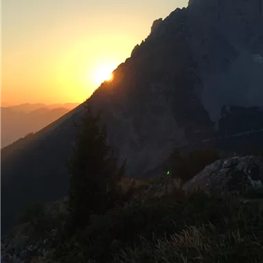 Ein Sonnenuntergang hinter einem majestätischen Berg. Die Landschaft ist von sanften Hügeln und Bäumen umgeben.