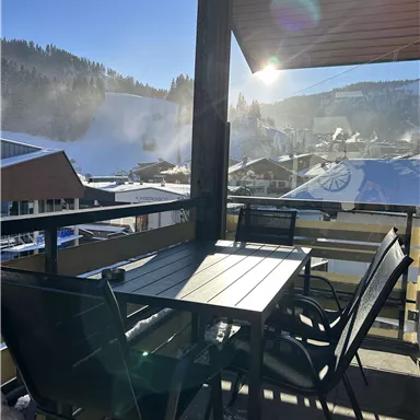 A balcony with a table and chairs that offers a view of snow-covered mountains. The sun shines brightly over the landscape.