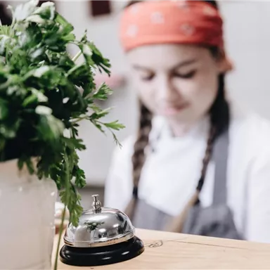 A cook with a red headscarf is working in a kitchen. In the foreground, there is a flower pot with fresh parsley.