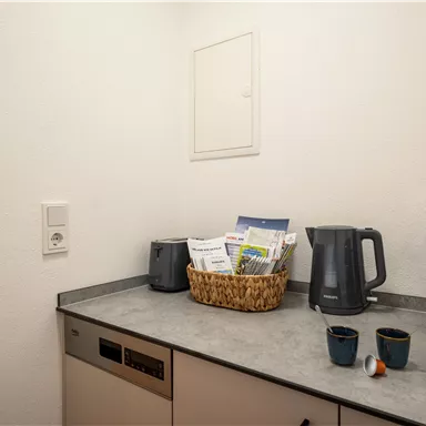A modern kitchen with a gray workspace. On the counter there is a kettle, a coffee capsule machine, and a basket with magazines.