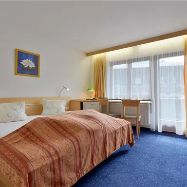 A cozy hotel room with two single beds and colorful bedding. The room has large windows that let in plenty of daylight.