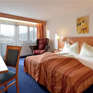 Ein komfortables Hotelzimmer mit einem Doppelbett, das mit Kissen dekoriert ist. Es gibt einen Schreibtisch, einen Stuhl und ein gemütliches Sessel neben einem Fenster mit Vorhängen.