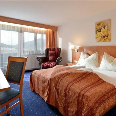 A comfortable hotel room with a double bed decorated with pillows. There is a desk, a chair, and a cozy armchair next to a window with curtains.