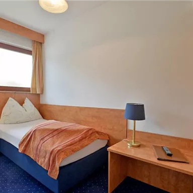 Ein einfaches Zimmer mit einem Einzelbett und Holzdekor. Es gibt einen Tisch mit einer Lampe und ein Fenster, das Tageslicht hereinlässt.