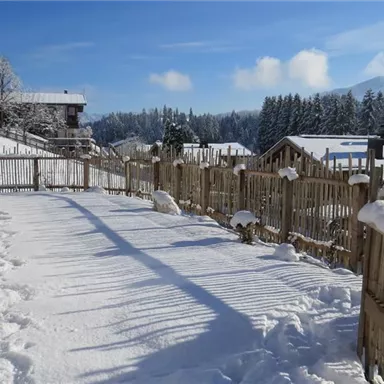 Eine verschneite Landschaft mit einem eingezäunten Bereich und Holzlattenzaun. Im Hintergrund sind Berge und ein klarer blauer Himmel zu sehen.