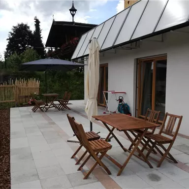 Eine gemütliche Terrasse mit Holzmöbeln und einem Sonnenschirm. Der Bereich ist von einem Zaun umgeben und hat eine grüne Umgebung.