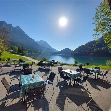 Eine schöne Terrasse mit Tischen und Stühlen am See. Im Hintergrund sind Berge und ein klarer Himmel zu sehen.