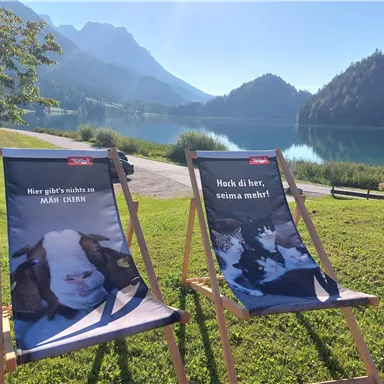 Zwei Liegestühle stehen am Ufer eines klaren Sees mit Bergen im Hintergrund. Auf den Stühlen stehen einladende Sprüche.
