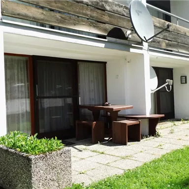Ein Außenbereich mit einem Tisch und Stühlen vor einem Gebäude. Die Terrasse ist von grüner Wiese umgeben und hat einen Satellitenspiegel.