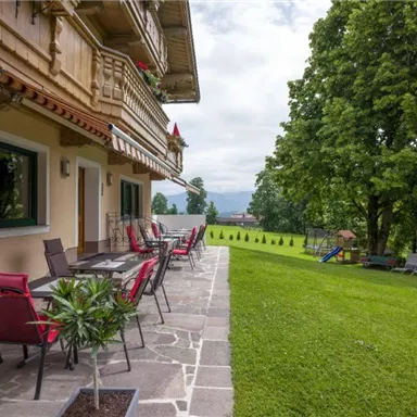 A terrace with red chairs and tables next to a well-kept garden. In the background, trees and a gentle landscape can be seen.