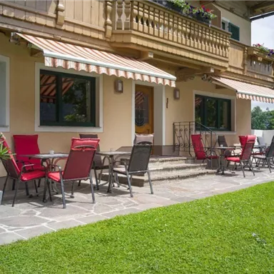 A beautiful terrace with red chairs and tables in front of a building. The surroundings are green and inviting.