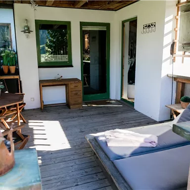 Eine gemütliche Terrasse mit einem Holzfußboden und mehreren Sitzgelegenheiten. Die Wände sind in hellen Farben gestrichen und die Fenster bieten einen Blick ins Grüne.