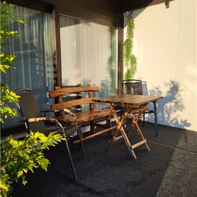 Ein gemütlicher Balkon mit einem Holztisch und zwei Stühlen. Im Hintergrund sind Pflanzen und Fenster zu sehen.