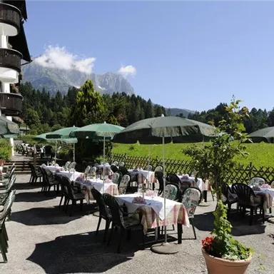 An inviting outdoor restaurant with covered tables and green umbrellas. In the background, there are blooming meadows and majestic mountains.
