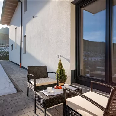 Eine gemütliche Terrasse mit modernen Gartenmöbeln und einer schönen Aussicht. Die Sonne scheint auf den schneebedeckten Boden.