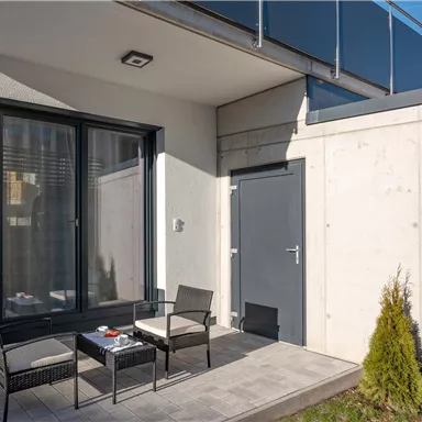 Eine moderne Terrasse mit zwei Stühlen und einem Tisch. Im Hintergrund befindet sich eine graue Wand und große Fenster.