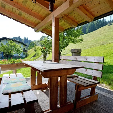 A cozy wooden porch with a table and benches. In the background, you can see a green meadow and gentle hills.