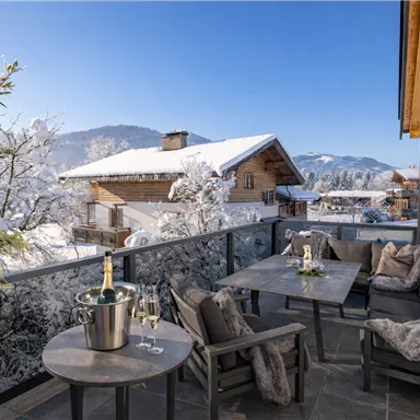 Eine gemütliche Terrasse mit einem Tisch und Stühlen, umgeben von schneebedeckten Bergen. Im Hintergrund sind traditionelle Holzhäuser und blauer Himmel zu sehen.