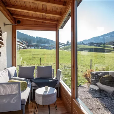 Ein gemütlicher Balkon mit einer Sitzgelegenheit und Blick auf grüne Wiesen und Berge. Die große Fensterfront sorgt für viel Tageslicht und einen schönen Ausblick.