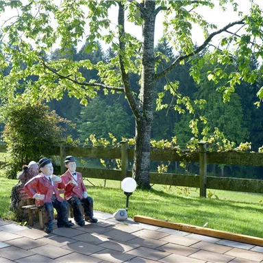 Three figures are sitting on a bench under a tree. In the background, a green meadow with a fence and trees is visible.