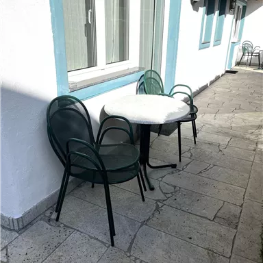 A small table with two chairs is set on a paved walkway. The wall is white with blue window frames.