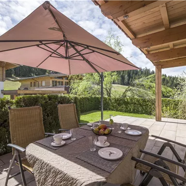A cozy terrace with a table, chairs, and a sunshade. In the background, there are green meadows and wooden buildings.