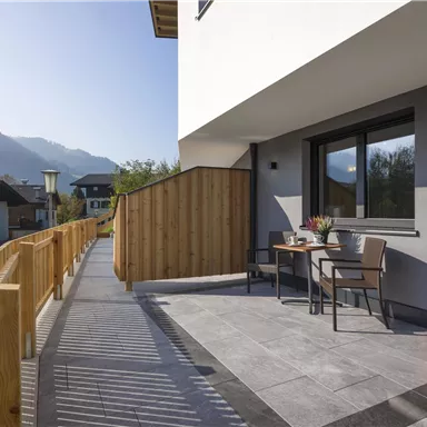 Eine moderne Terrasse mit einem Tisch und zwei Stühlen. Im Hintergrund sind Berge und Häuser zu sehen.