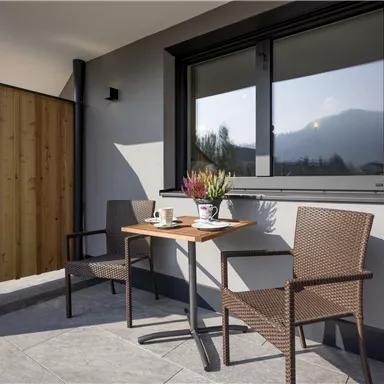 Ein moderner Balkon mit einem kleinen Tisch und zwei Stühlen. Auf dem Tisch stehen eine Tasse und eine Blumenvase.