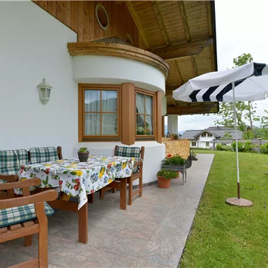 A cozy terrace with a decorative table and chairs. A parasol protects from the sun, surrounded by green grass and plants.