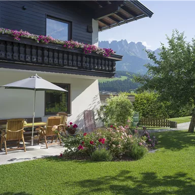A pretty house with a balcony adorned with blooming flowers. In front of the house is a cozy garden with a terrace and a view of the mountains.