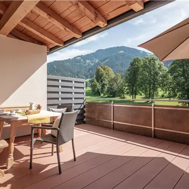 A cozy balcony with wooden furniture and a table. In the background, green meadows and mountains can be seen.