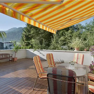 Eine gemütliche Terrasse mit einem Tisch und Stühlen. Im Hintergrund sind Berge und Pflanzen zu sehen, dazu ein sonnenschützendes, gestreiftes Sonnensegel.