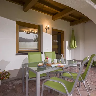 Ein einladender Patio mit einem Glas-Tisch und grünen Stühlen. Im Hintergrund sieht man Fenster und eine grüne Markise.