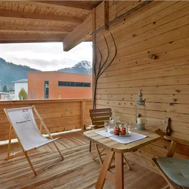 Ein gemütlicher Balkon aus Holz mit einem Tisch und zwei Stühlen. Im Hintergrund sind Berge und ein klarer Himmel zu sehen.