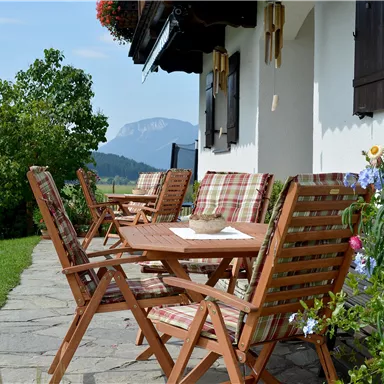 An inviting terrace with wooden chairs and a table, surrounded by greenery and flowers. In the background, mountains and a clear sky can be seen.