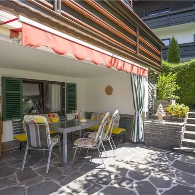 Eine gemütliche Terrasse mit einem Esstisch und Stühlen, umgeben von Pflanzen. Der Bereich hat einen sonnigen und einladenden Charakter.