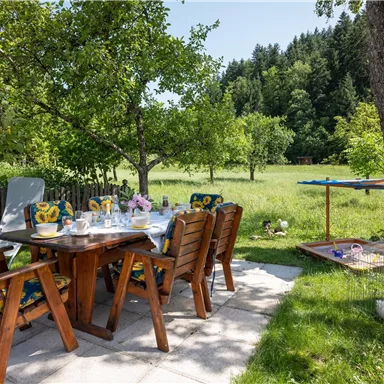 An inviting outdoor dining area with wooden furniture and blooming plants. In the background, a green meadow stretches out with trees.