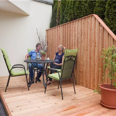 Ein gemütlicher Patio mit Holzterrasse und grünen Stühlen. Zwei Personen sitzen entspannt am Tisch und genießen ihre Getränke.