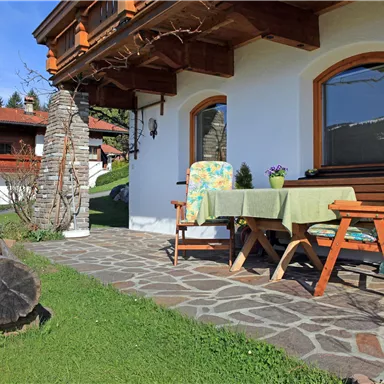 Ein gemütlicher Außenbereich mit einem Tisch und Stühlen. Im Hintergrund sind grüne Wiesen und ein traditionelles Haus zu sehen.