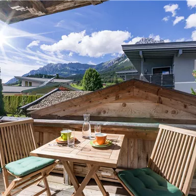 Eine gemütliche Terrasse mit Blick auf die Berge. Zwei Stühle und ein Tisch sind mit Getränken dekoriert, während die Sonne strahlt.