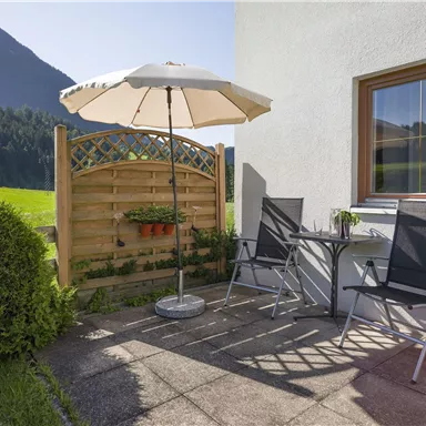 Eine gemütliche Terrasse mit zwei Stühlen und einem Tisch. Ein Sonnenschirm bietet Schatten, und im Hintergrund sind grüne Wiesen und Berge zu sehen.