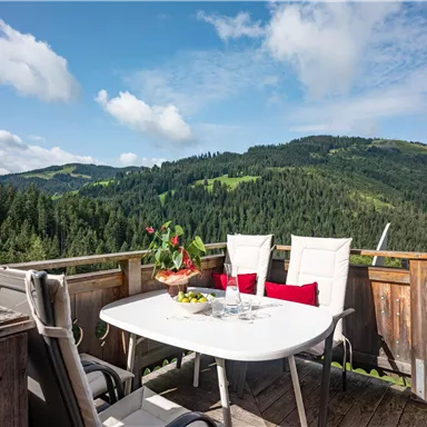 Ein schöner Balkon mit einem Tisch und Stühlen, umgeben von einer beeindruckenden Berglandschaft. Die Aussicht zeigt grüne Wälder und sanfte Hügel unter einem blauen Himmel.