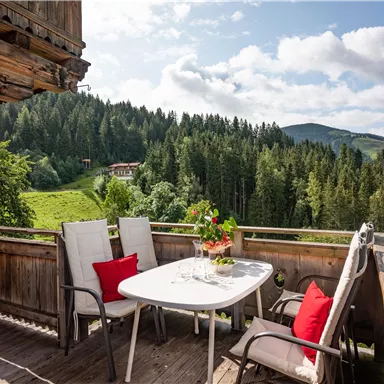 Ein gemütlicher Balkon mit einem Tisch und Stühlen, umgeben von üppigem Grün und bewaldeten Hügeln. Die Aussicht ist malerisch und es gibt einen blauen Himmel mit einigen Wolken.