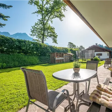 Ein schöner Garten mit einer Terrasse und einem Tisch. Im Hintergrund sind Bäume und Berge sichtbar, der Himmel ist klar und sonnig.