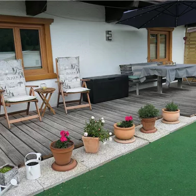 A cozy terrace with two deck chairs, a table, and a sun umbrella. Flower pots and a green lawn create an inviting atmosphere.