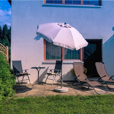 Ein gemütlicher Außenbereich mit Liegestühlen und einem Sonnenschirm. Der Bereich ist umgeben von einer grünen Rasenfläche und bietet eine entspannte Atmosphäre.