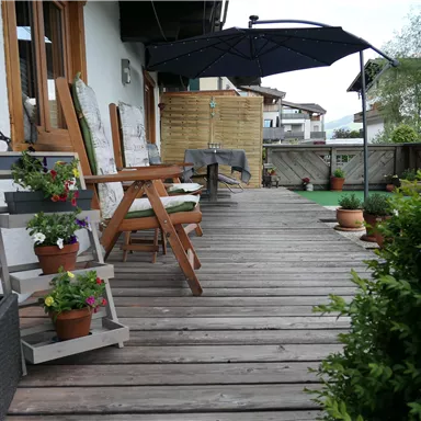 A cozy terrace with wooden planks, chairs, and blooming plants. A sun umbrella provides shade and there is a view of the surroundings.