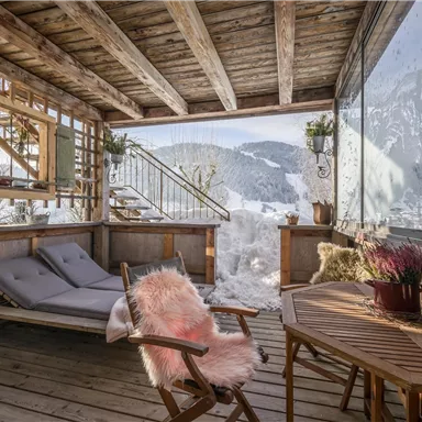 Ein gemütlicher Wintergarten mit Holzverkleidung und Blick auf verschneite Berge. Ein bequemes Sofa und ein Holztisch laden zum Entspannen ein.